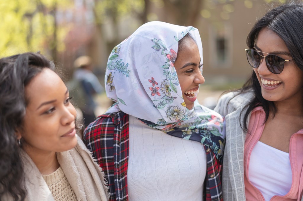 A small group of university women poses outdoor on their university campus. They are smiling as they discuss their test results in their most recent class. They are diverse in ethnic origin.