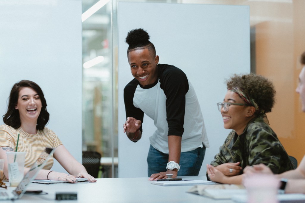 A multiracial group of college students in their twenties laugh together while meeting in a library study room.