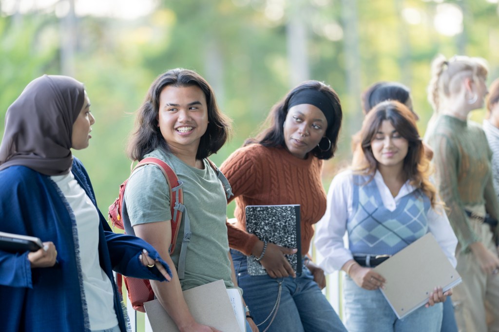 A large group of University students casually stand beside each other along a railing, as as they pose together for a portrait.  They are each dressed casually and are smiling as they hold their books and chat.