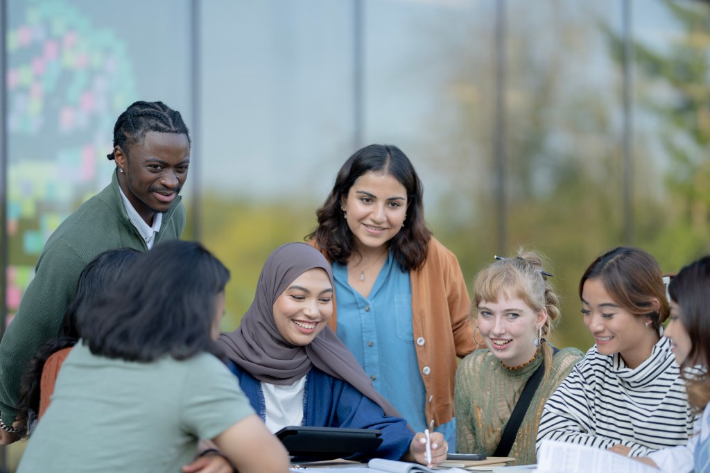 A small group of University students sit around a table outside on campus as they work on a group assignment.  They have textbooks spread out in front of them and are using a tablet to reference material as they work together.
